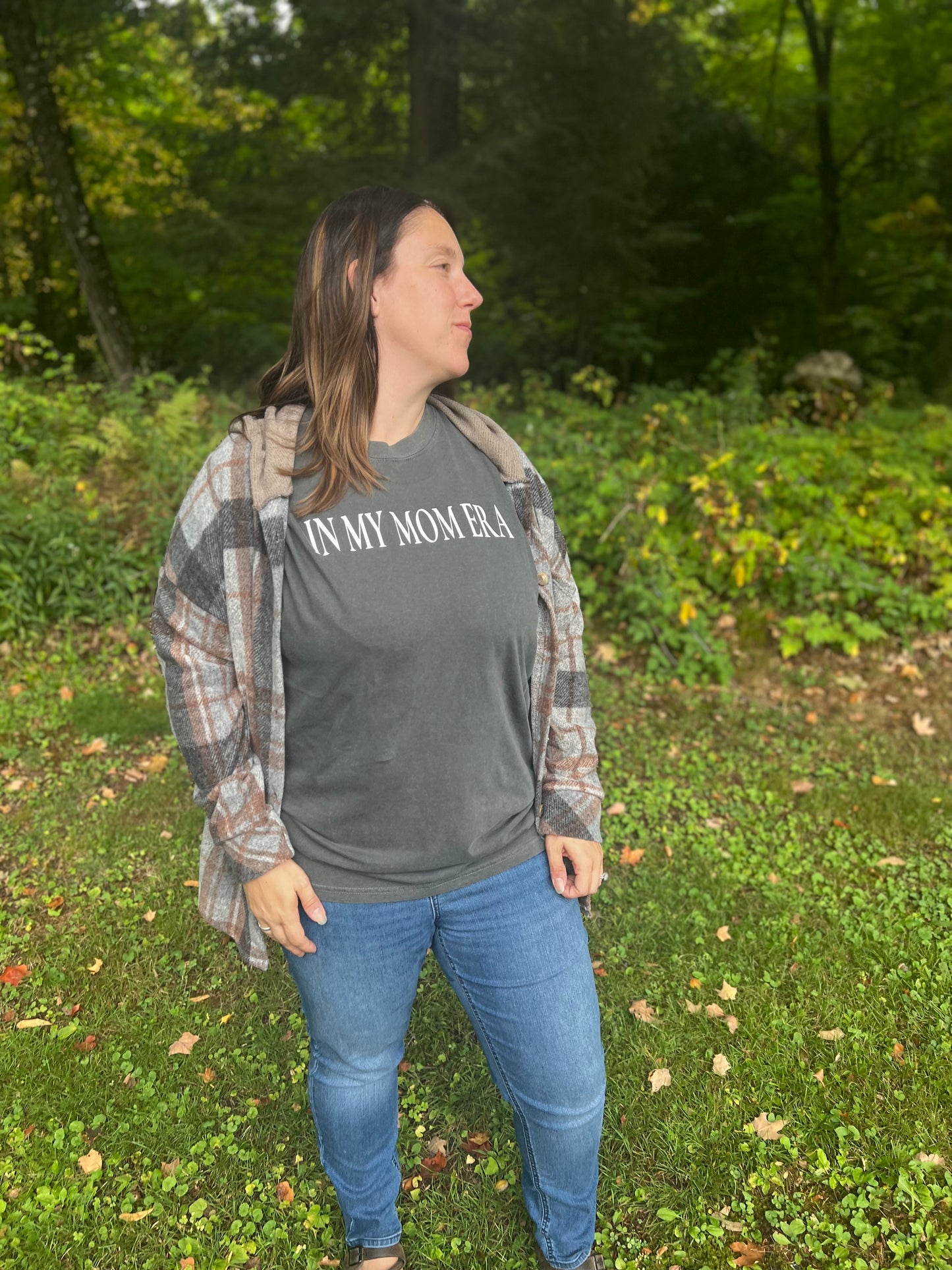 A mid-size woman in her thirties modeling a charcoal graphic tee with saying, "In My Mom Era." She also is wearing a orange and charcoal plaid hoodie shacket.